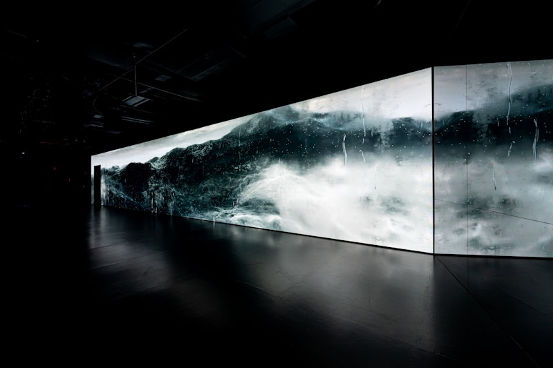 En stor skærm viser en dynamisk, stormfuld havscene med voldsomme bølger i et mørkt, minimalistisk rum.