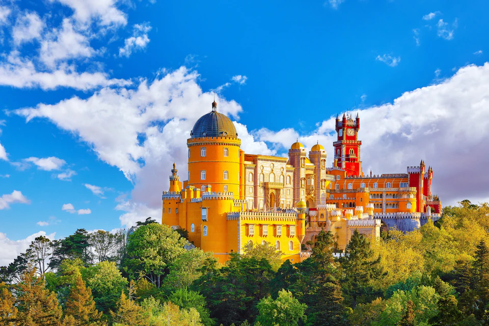 A vibrant yellow and red palace with domes and towers, surrounded by green trees, under a bright blue sky with clouds.