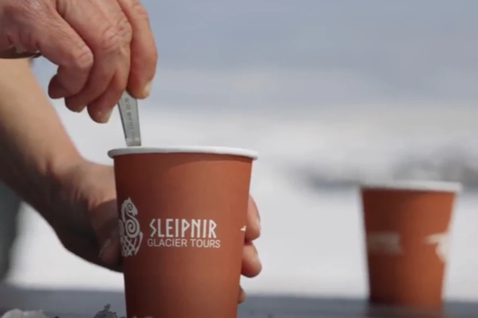A hand stirs a drink in an orange cup labeled "Sleipnir Glacier Tours" with an ocean background.