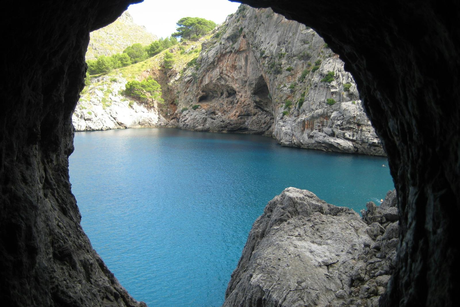 View from a cave overlooking a serene, blue body of water surrounded by rocky cliffs and greenery.