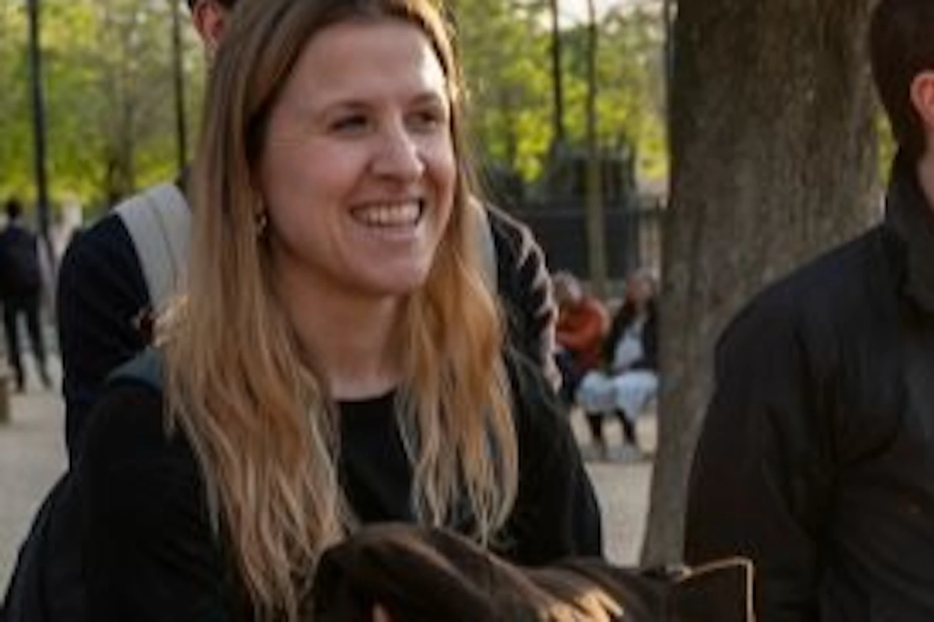 A group of people talking and smiling in a park with trees, sunlight filtering through, and a distant background of benches.