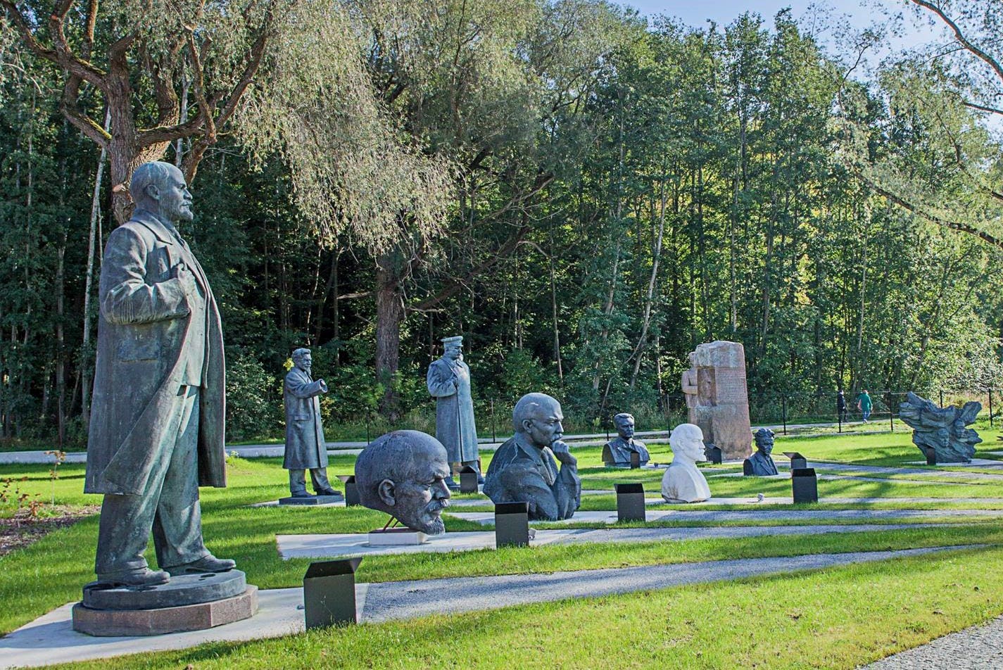 Parque ao ar livre com várias estátuas e bustos de figuras históricas dispostas num relvado relvado com árvores ao fundo.
