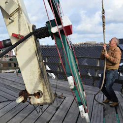 En man i en brun jacka drar ett tjockt rep på ett trädäck bredvid stora mekaniska apparater med byggnader i bakgrunden.