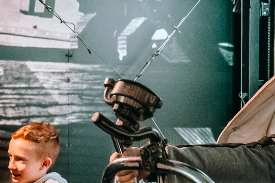 A child sits on a decorative steampunk-style flying bicycle exhibit, smiling and giving a thumbs-up, with another child nearby.