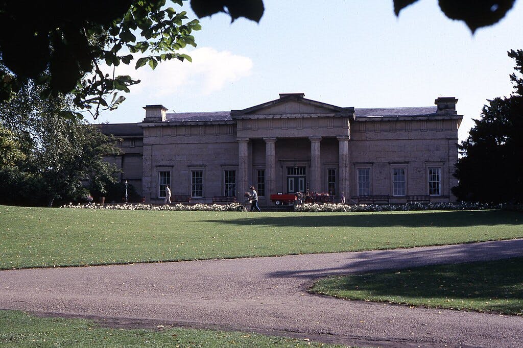 Neoklasicistní budova se sloupy, dvěma procházejícími se lidmi, červeným vozidlem a okolním zeleným trávníkem a stromy.