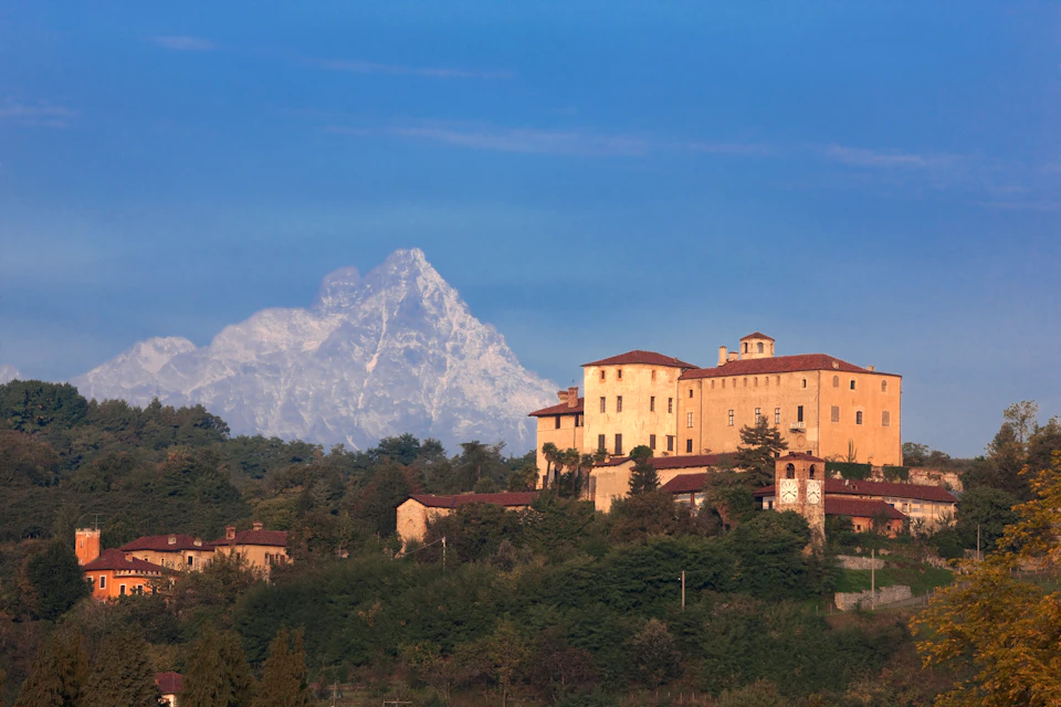 Historic buildings on a hill with a forested area in the foreground and a snow-capped mountain in the background under a clear sky.