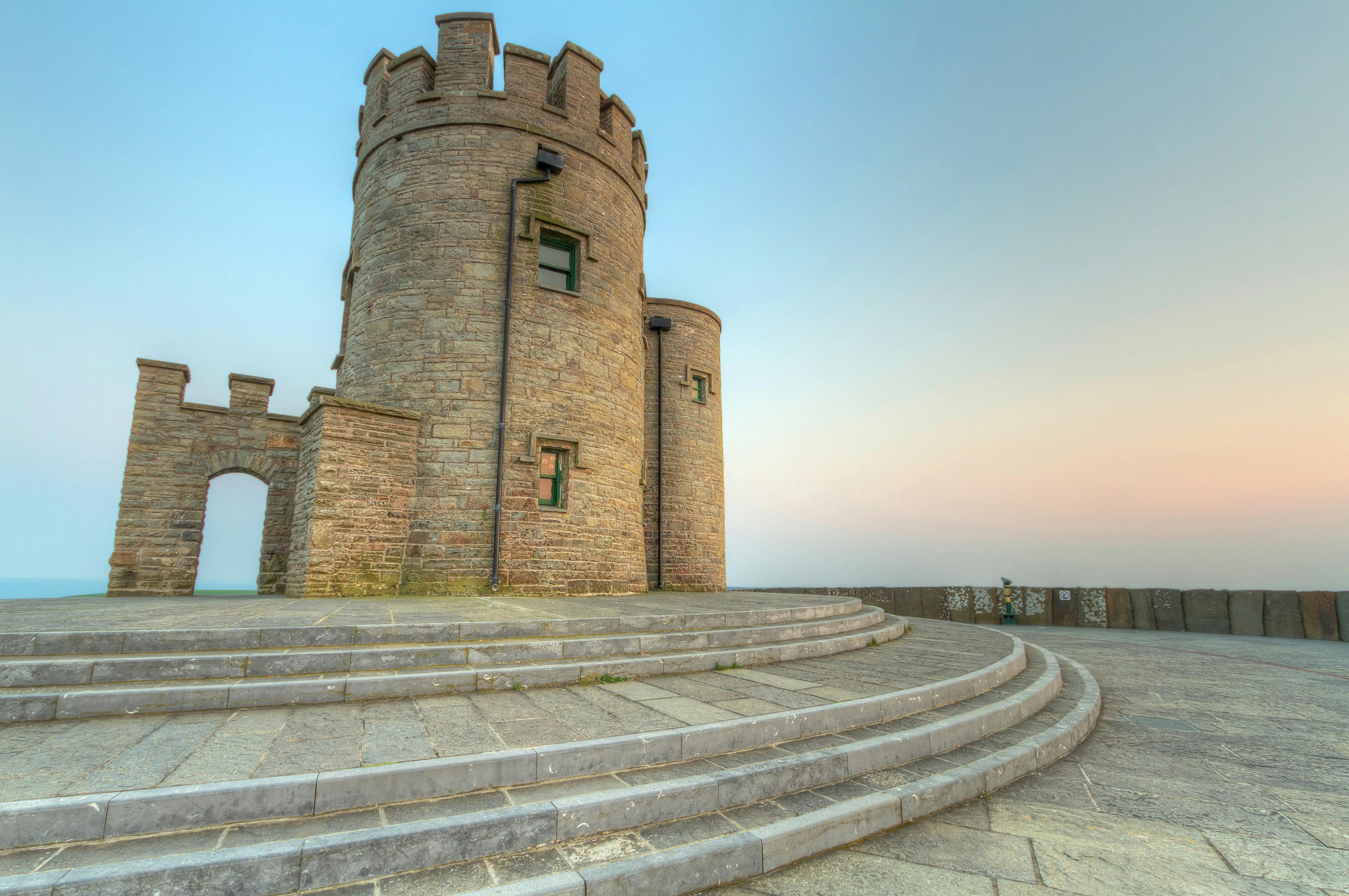 A round stone tower with small windows and battlements, surrounded by curved stone steps, under a clear sky at dusk.