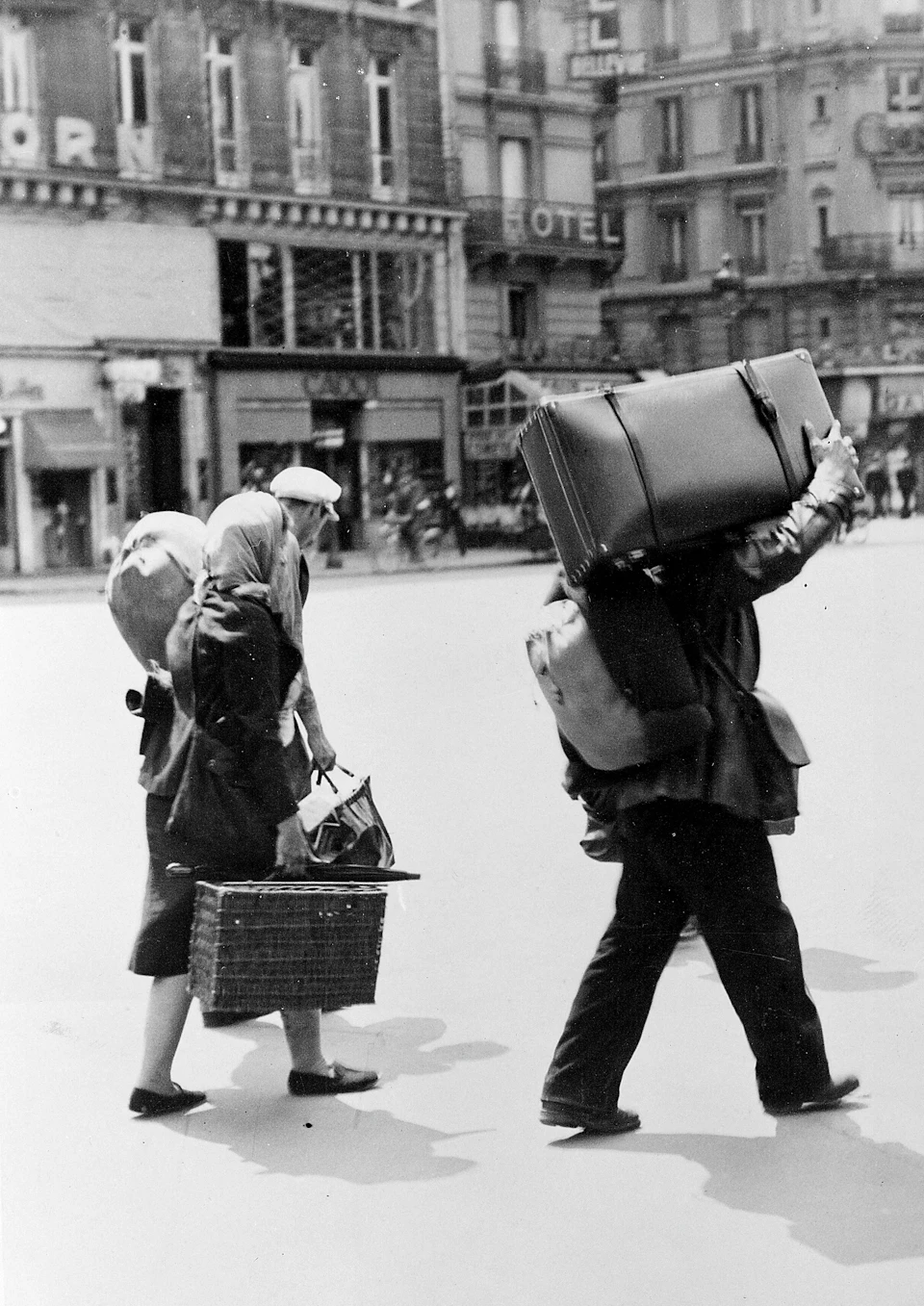 Two people crossing a street, carrying large bags and a suitcase on their heads, with a backdrop of buildings and storefronts.