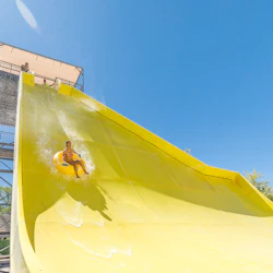 En person på ett gult rör glider nerför en brant gul vattenrutschbana under en klarblå himmel.