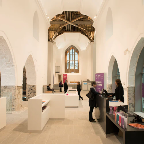 People interact and browse inside a modern museum with arched stone walls, informational displays, and a large arched window.