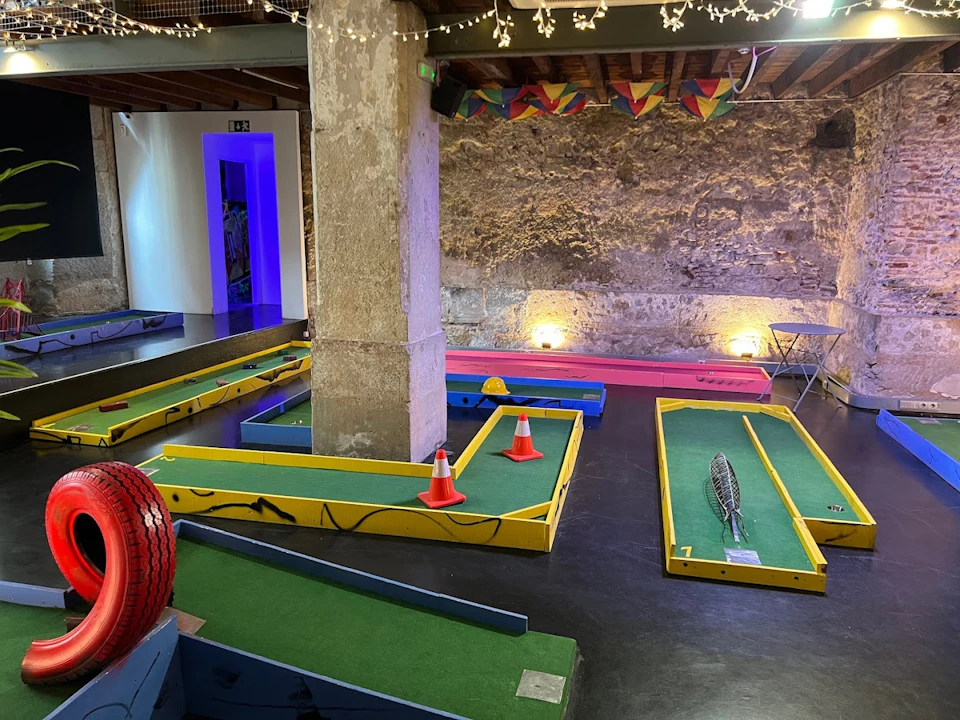Indoor mini-golf course with green turf, blue and yellow barriers, orange cones, a racket, and string lights overhead.