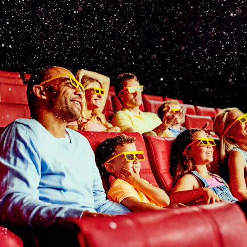 Människor som bär 3D-glasögon sitter i röda biosäten, ler och tittar på en film, med partiklar som flyter i luften.