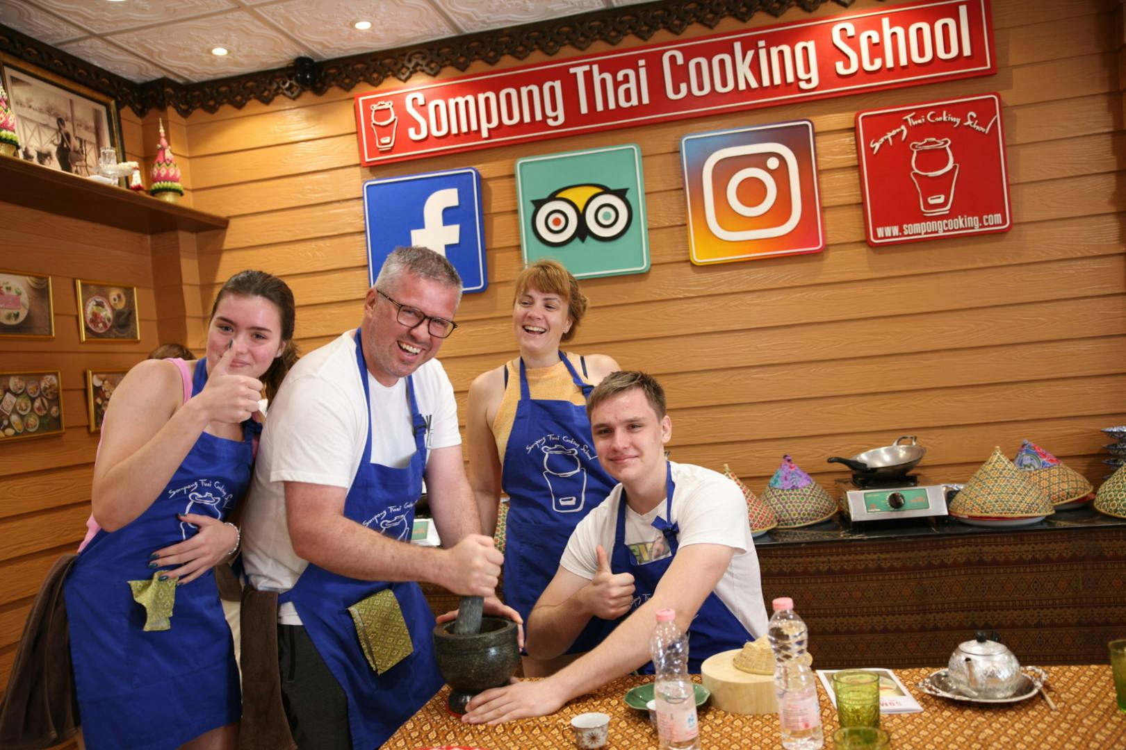 A group of four people wearing aprons participate in a cooking class at "Sompong Thai Cooking" with a mortar and pestle on a table.