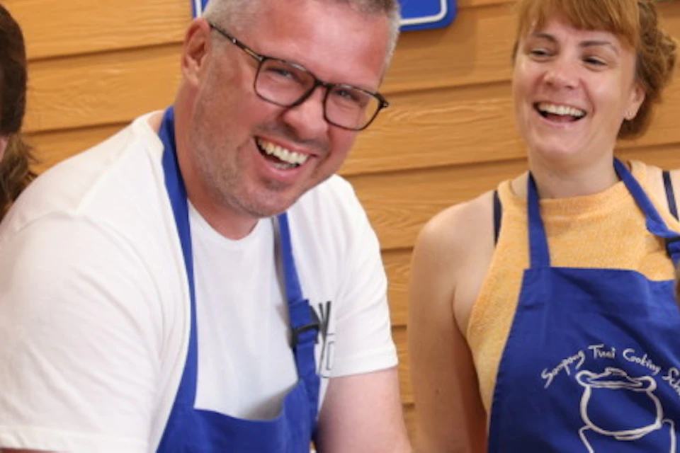 Four people wearing blue aprons at a cooking class, smiling and giving thumbs up, with a mortar and pestle on the table.