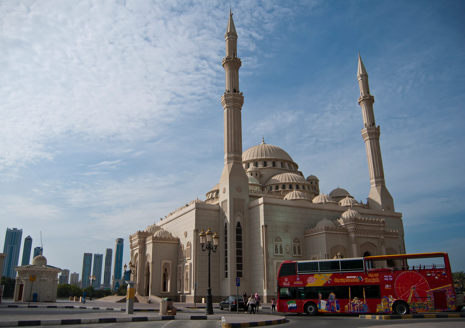 A large mosque with minarets under a partly cloudy sky, with a red double-decker bus and a few people nearby.
