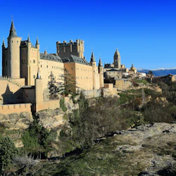 Ett storslaget medeltida slott med torn och befästningar ligger på en stenig kulle, med ett avlägset snötäckt berg och klarblå himmel.