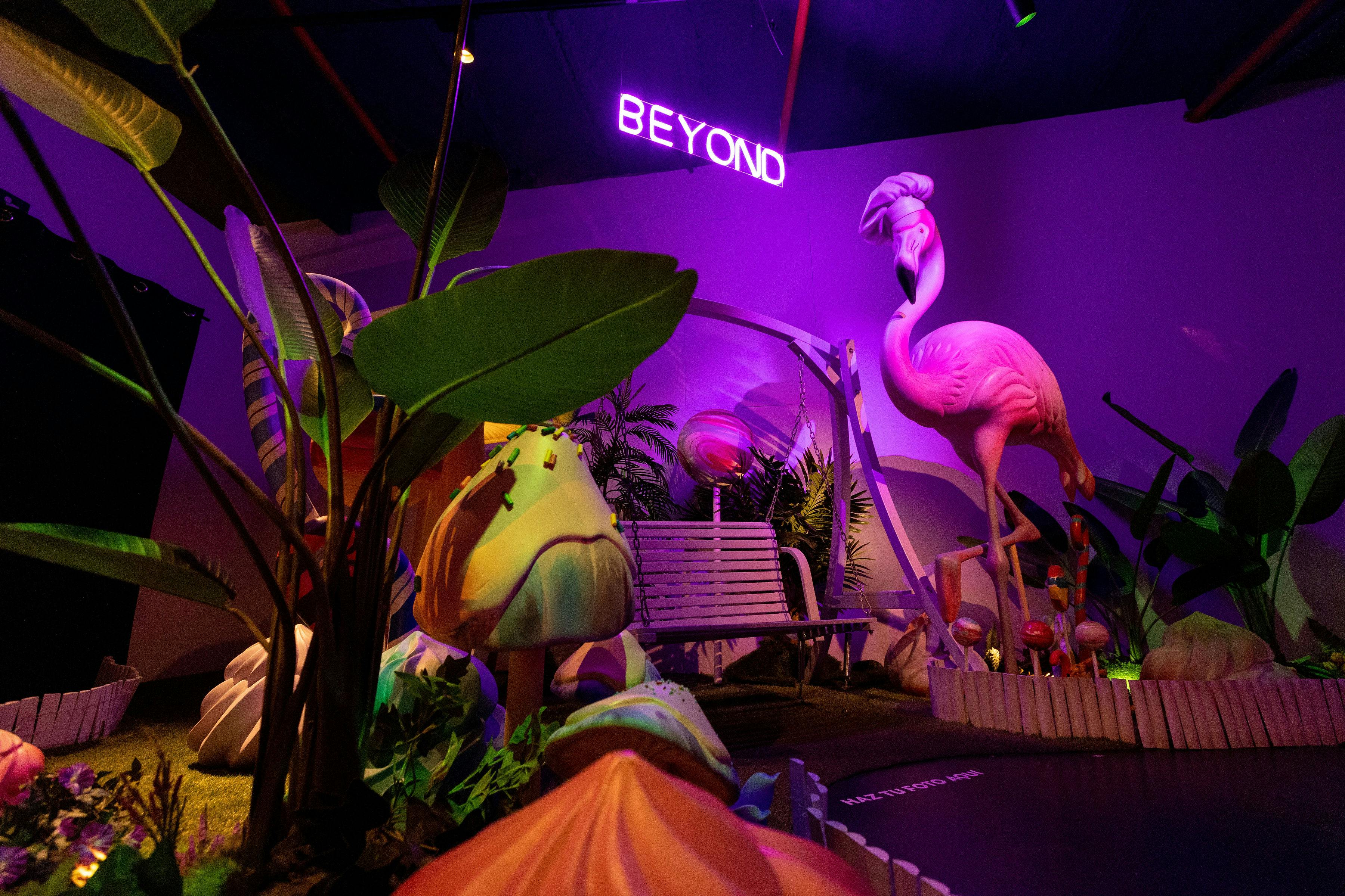 A brightly lit room with a large pink flamingo sculpture, neon &#34;BEYOND&#34; sign, and large leaves and mushrooms creating a whimsical environment.