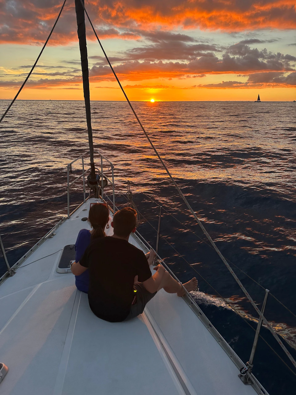 Two people sit on a sailboat's bow, watching a sunset over calm ocean waters, with another sailboat in the distance.