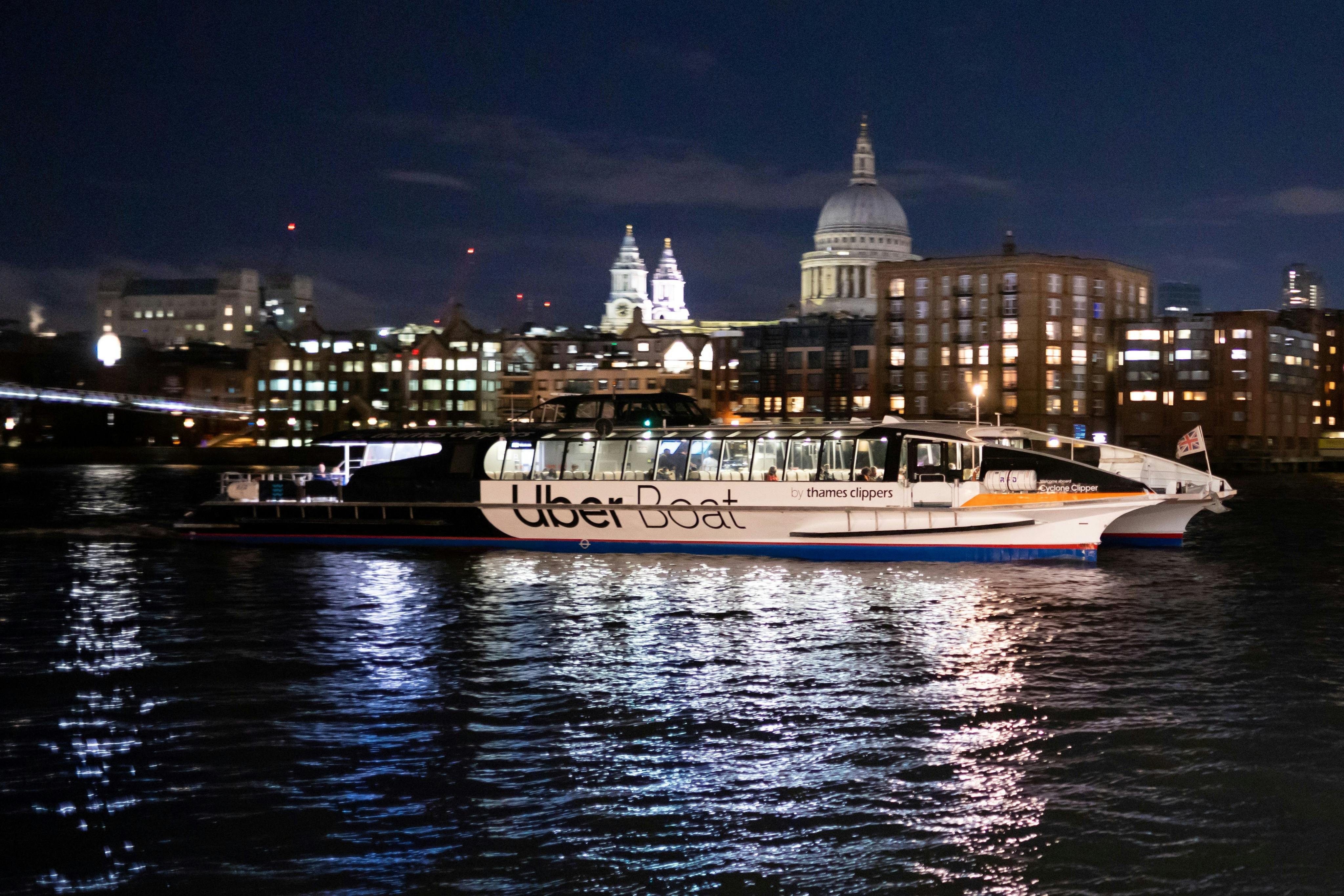 A boat labeled 'Uber Boat by Thames Clippers' docked on a river at night, with illuminated buildings and a dome in the background.