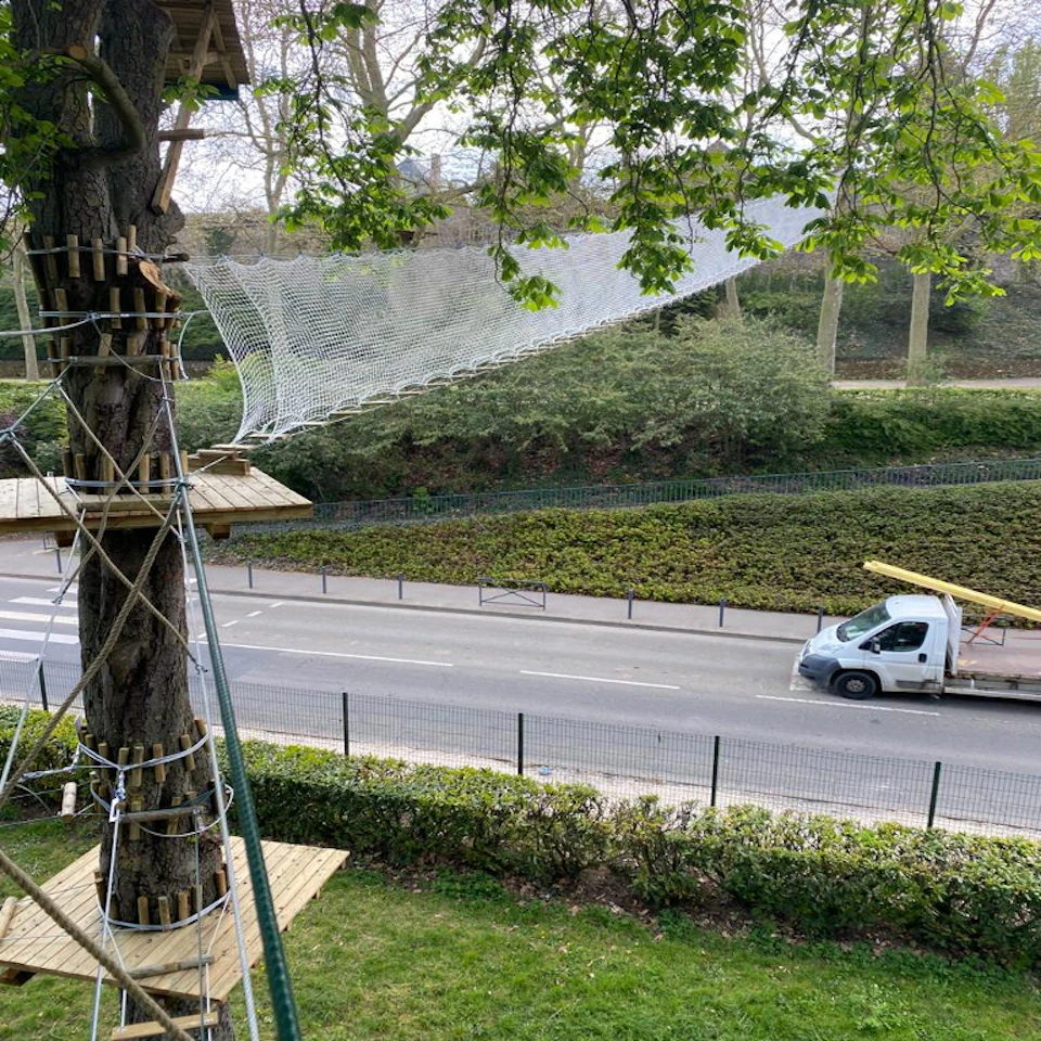 Wooden platforms connected by rope and nets are built around tree trunks. A road with a parked truck is visible below.