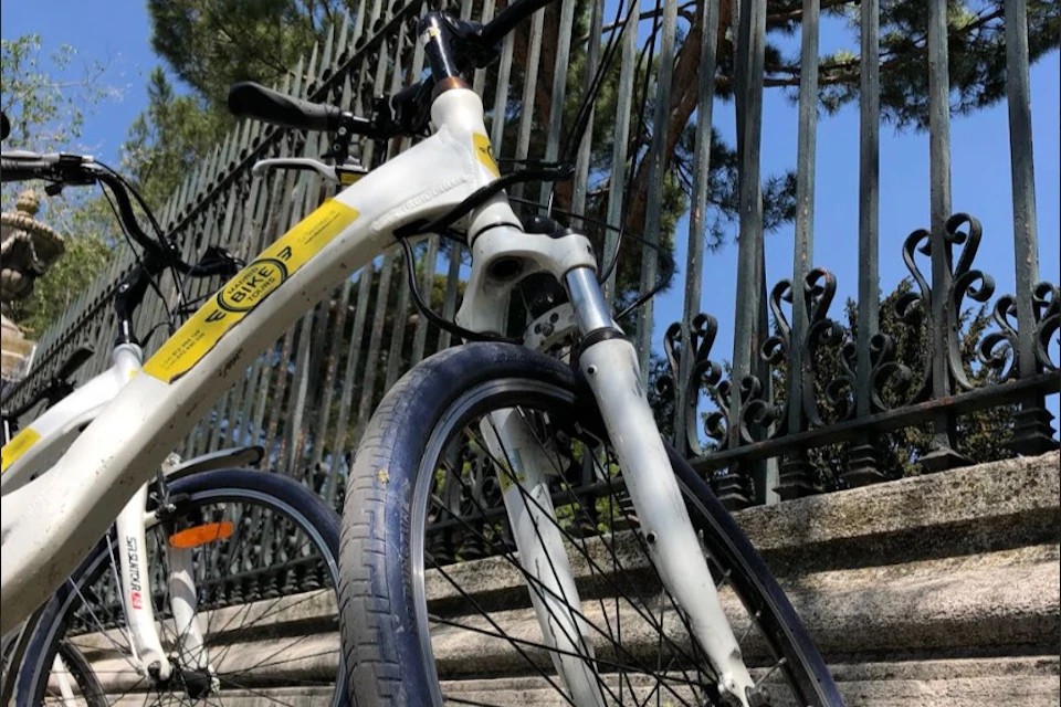 Close-up of a white bicycle with a yellow sticker parked beside a metal fence with ornate details. Trees are visible in the background.