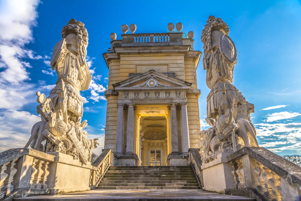 A grand stone building with columns, flanked by large detailed statues under a clear blue sky. Steps lead up to the entrance.