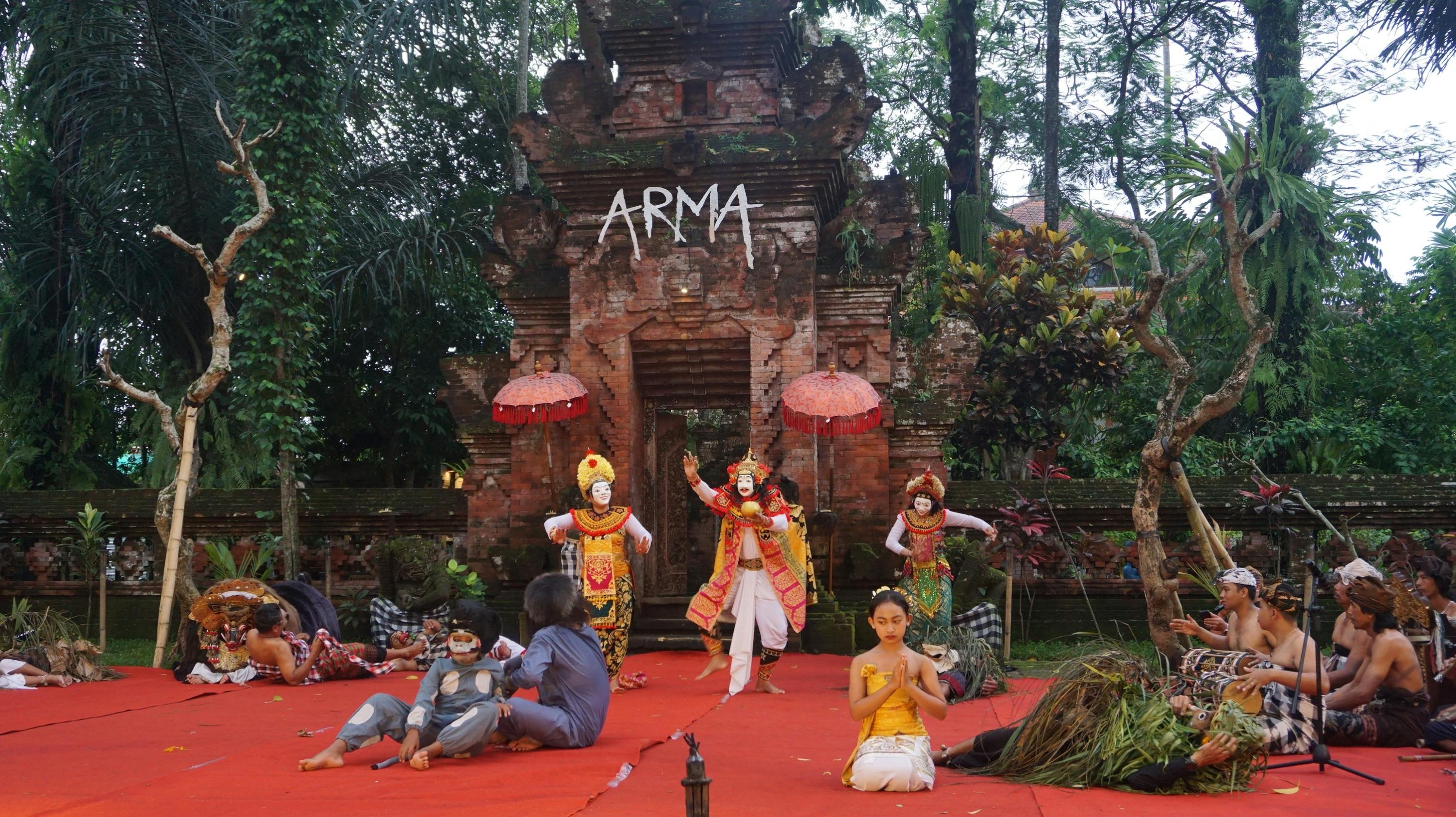 A traditional dance performance is taking place on a stage with ornate costumes, a brick structure in the background, and trees surrounding.