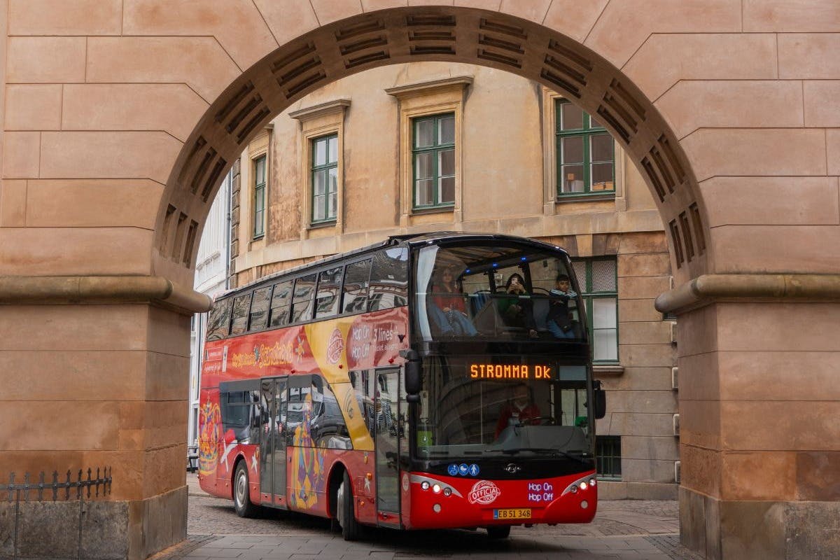 En röd dubbeldäckad turnébuss märkt "Stromma DK" kör genom en välvd stenport i en urban miljö.