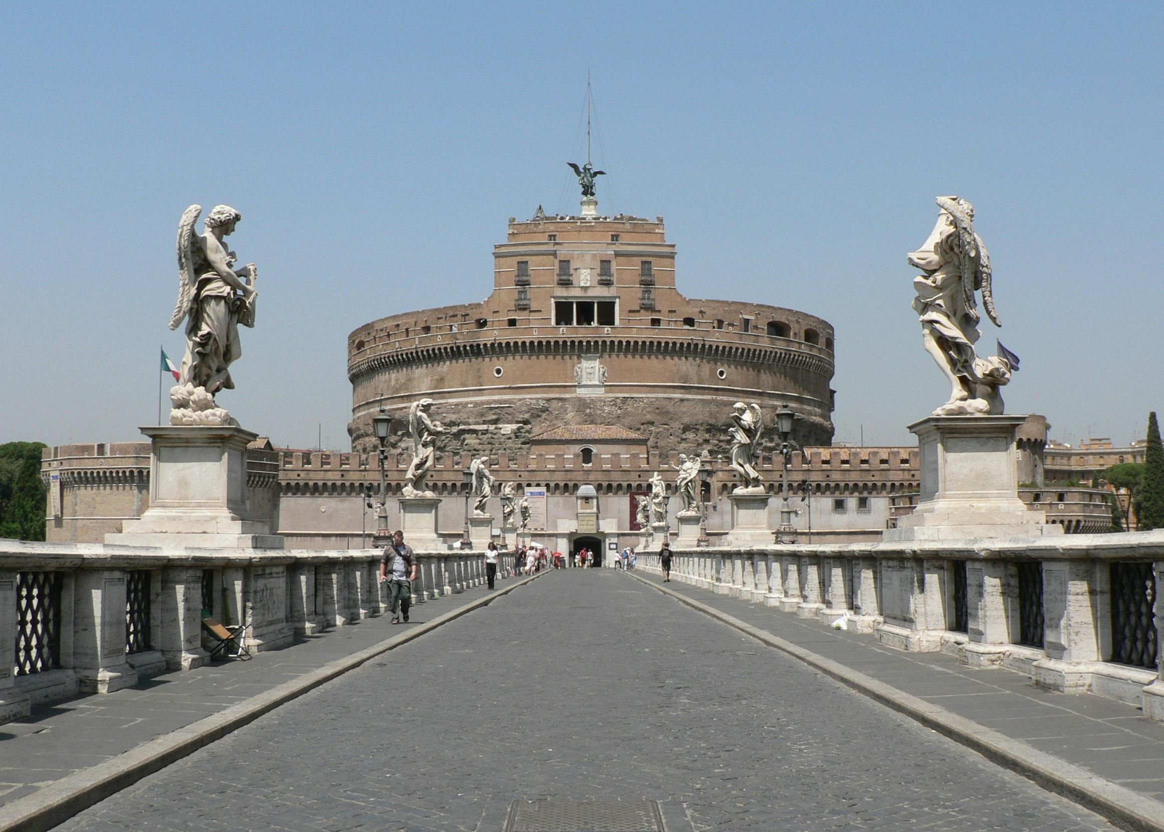 Wide stone bridge with statues leads to a large cylindrical castle topped with a statue, set against a clear blue sky.