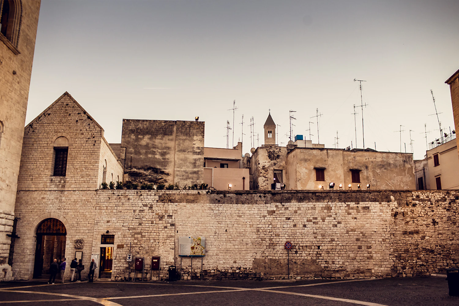 Old stone buildings with various antennas and a church tower in the background. A few people are seen near the entrance.