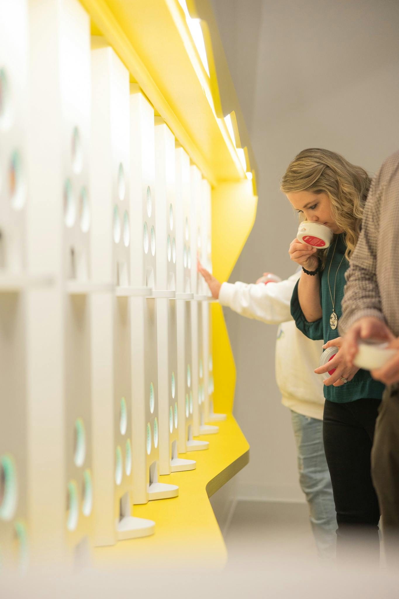 En kvinde nipper til en kop, mens hun står ved siden af en række hvide hylder med udskæringer og gul belysning. To andre personer er delvist synlige.