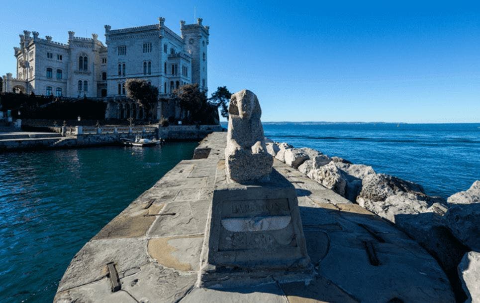 A stone statue on a pier with a castle-like building in the background, overlooking calm blue water under a clear sky.