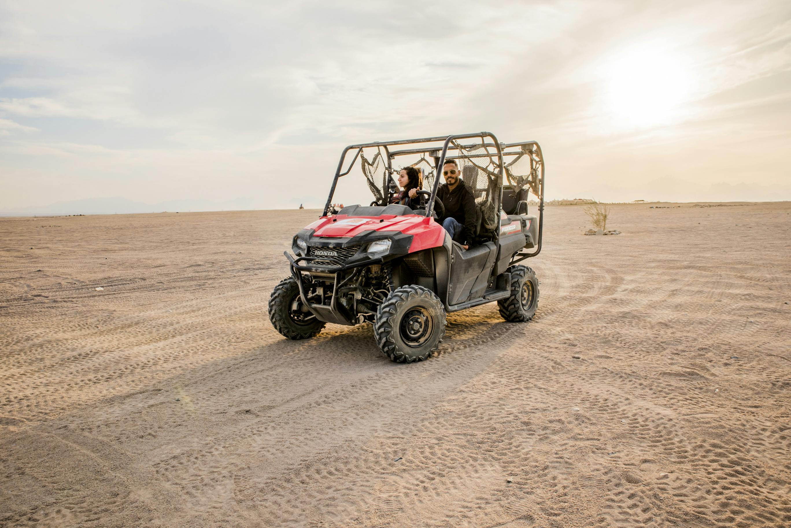 Dues persones conduint un vehicle tot terreny vermell i negre en un vast desert de sorra sota un cel parcialment ennuvolat.