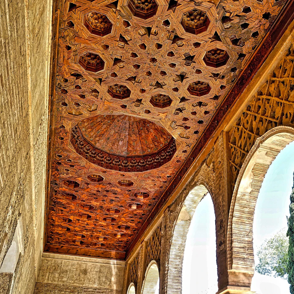 Intricately carved wooden ceiling and arched windows in a historic, sunlit corridor.
