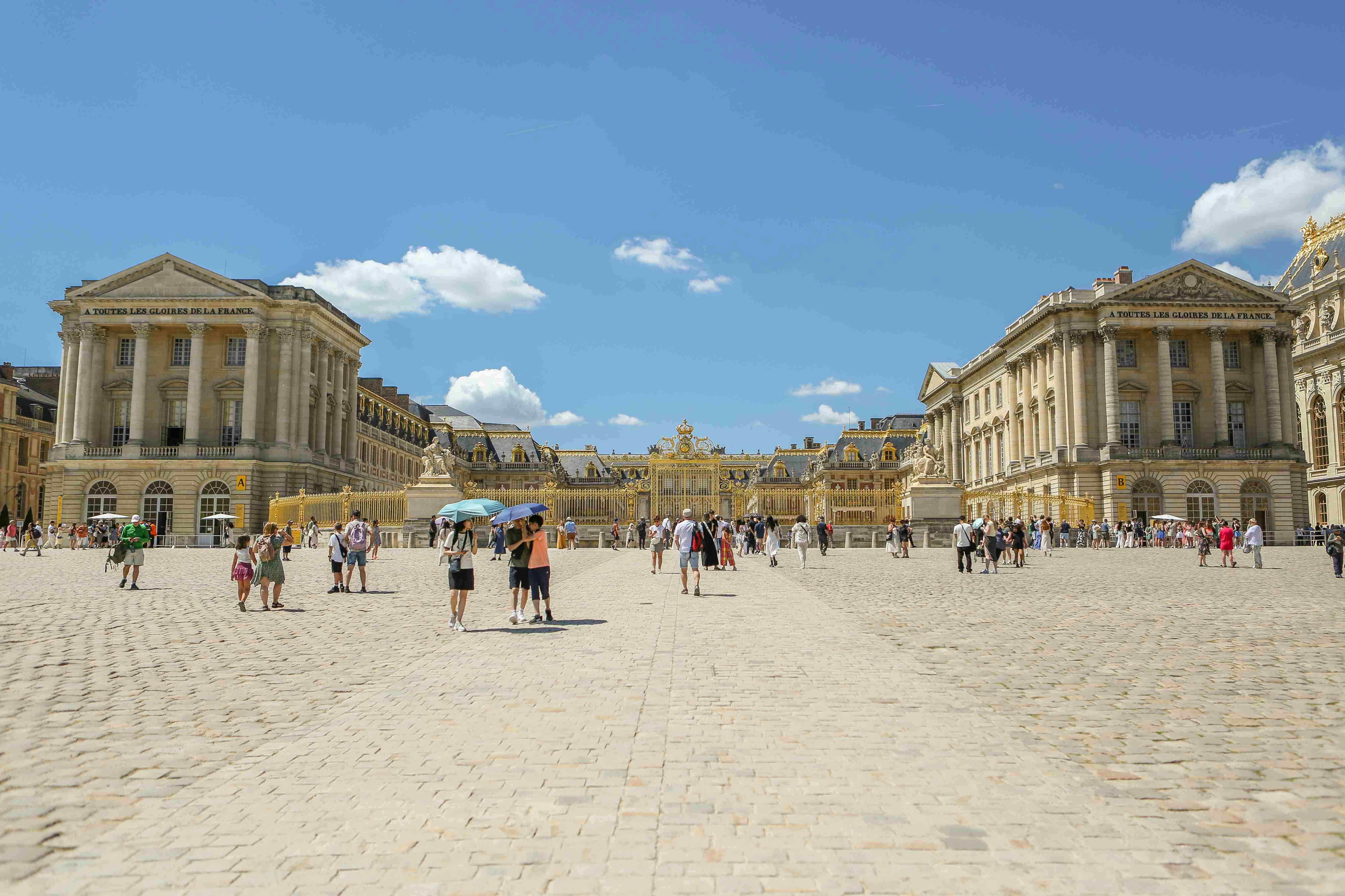Visitors walking on a wide cobblestone courtyard towards ornate, golden gates, flanked by symmetrical classical buildings under a blue sky.