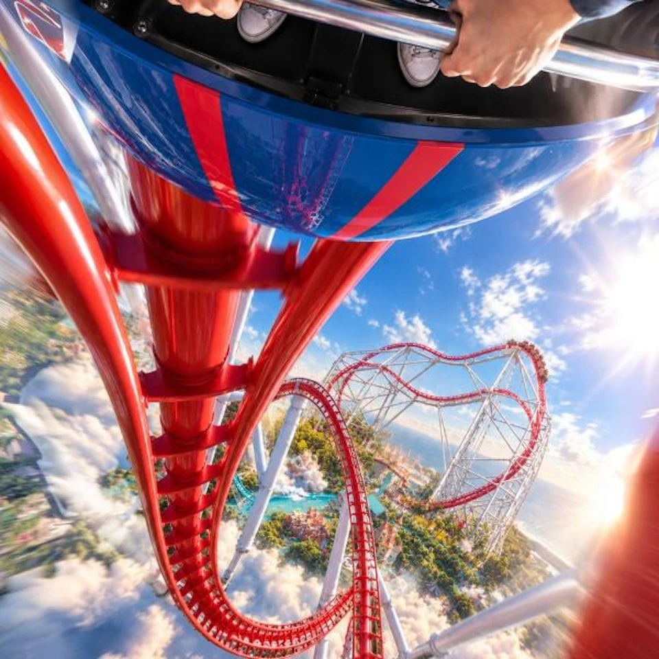 A first-person view from a rollercoaster descending a steep red track with twists, under a bright sky and scenic landscape.
