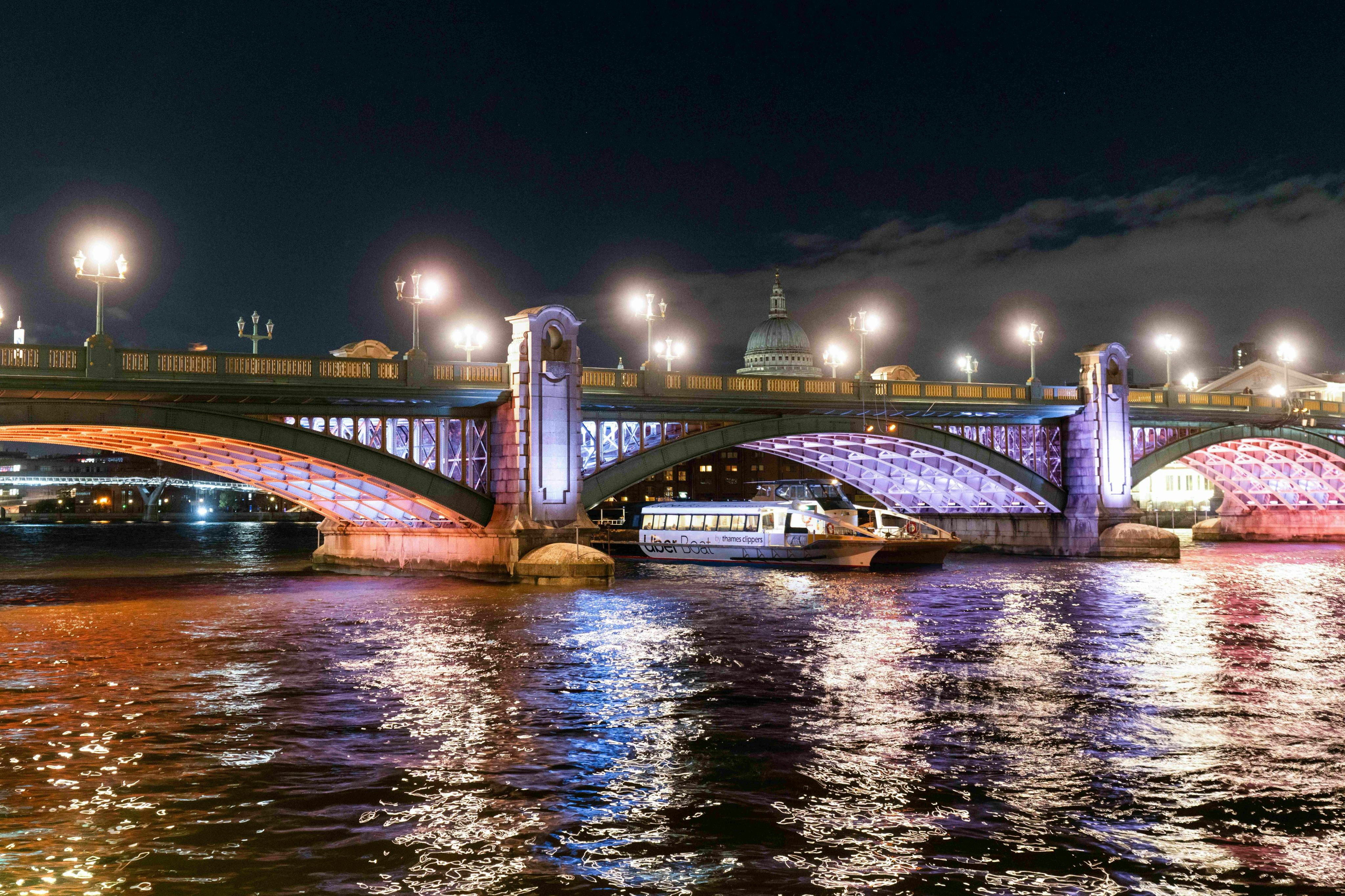 A night view of a bridge with decorative lighting over a river, with a boat passing underneath and a dome visible in the background.