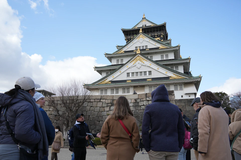 A group of people standing near Osaka Castle, a traditional Japanese castle with ornate roofing, under a partly cloudy sky.