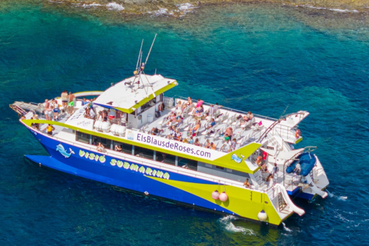 Un bateau d'excursion portant l'inscription « Els Blaus de Roses », avec des passagers à bord, naviguant dans des eaux d'un bleu limpide près d'un littoral rocheux.