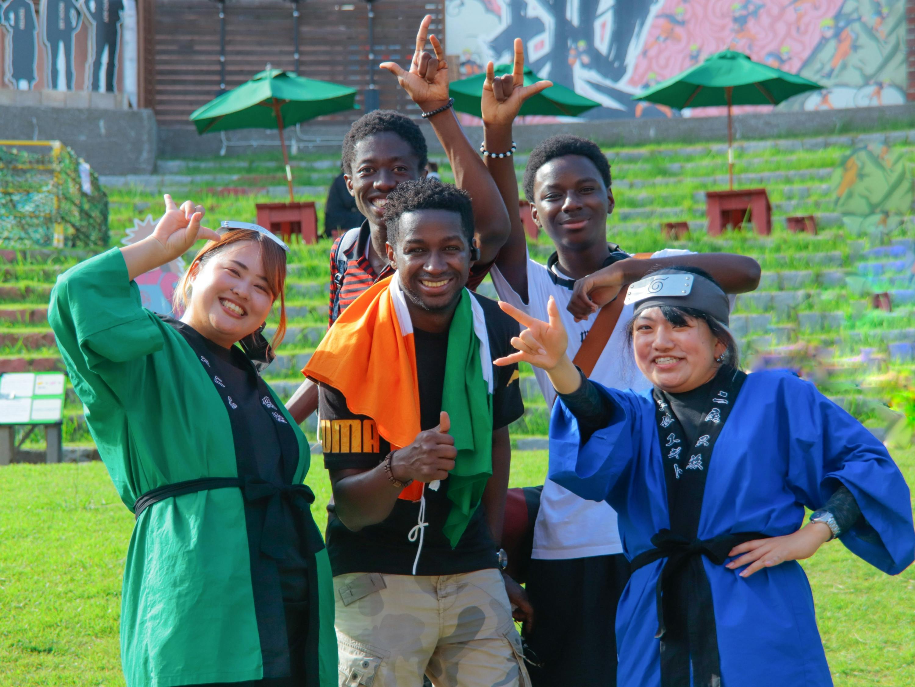 A group of five people smiling and posing outdoors in colorful attire, with a grassy area and umbrellas in the background.