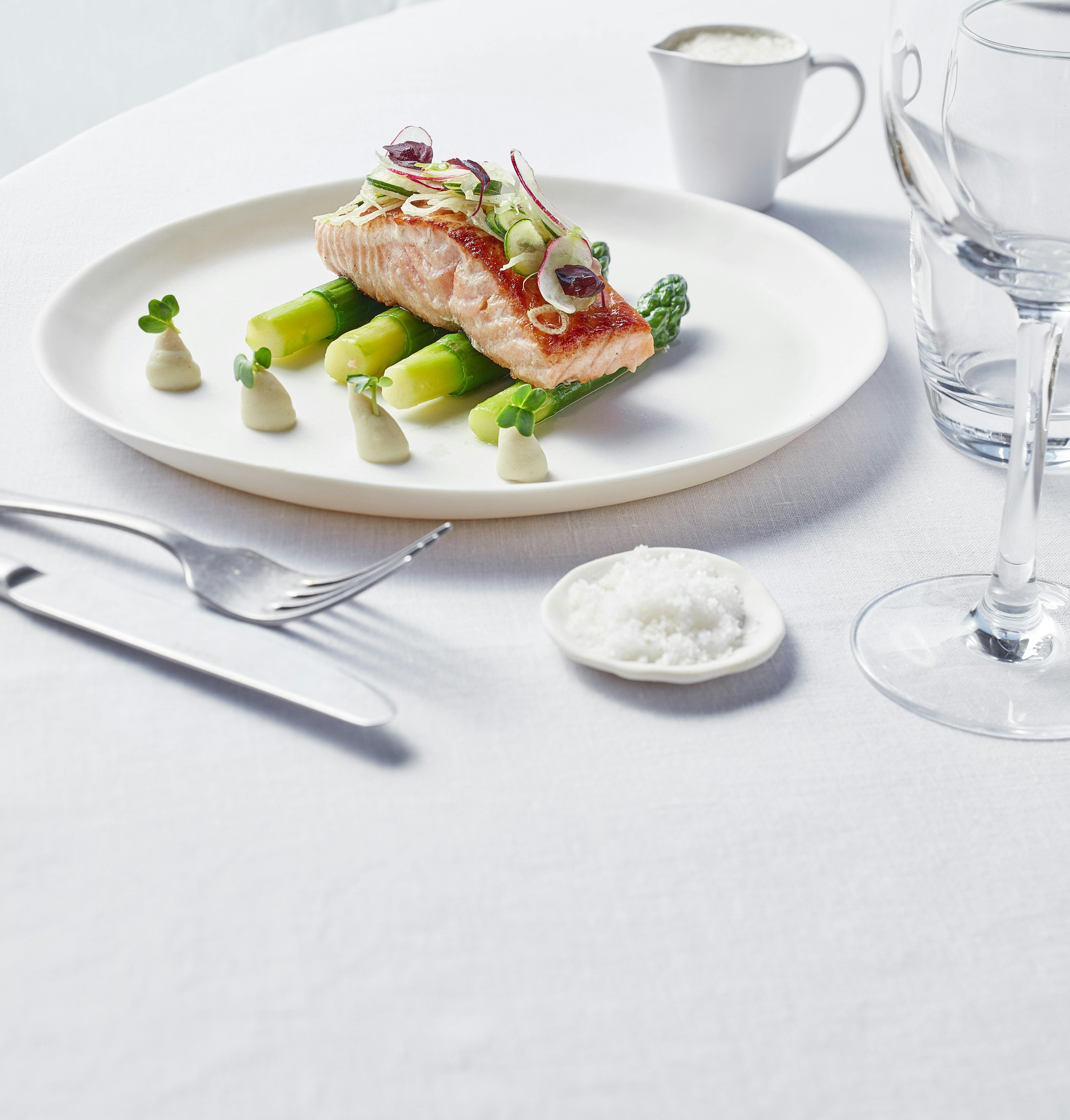 Filet de salmó sobre espàrrecs amb guarnicions, servit en un plat blanc amb estris, un got i un plat petit de sal sobre un estovall blanc.