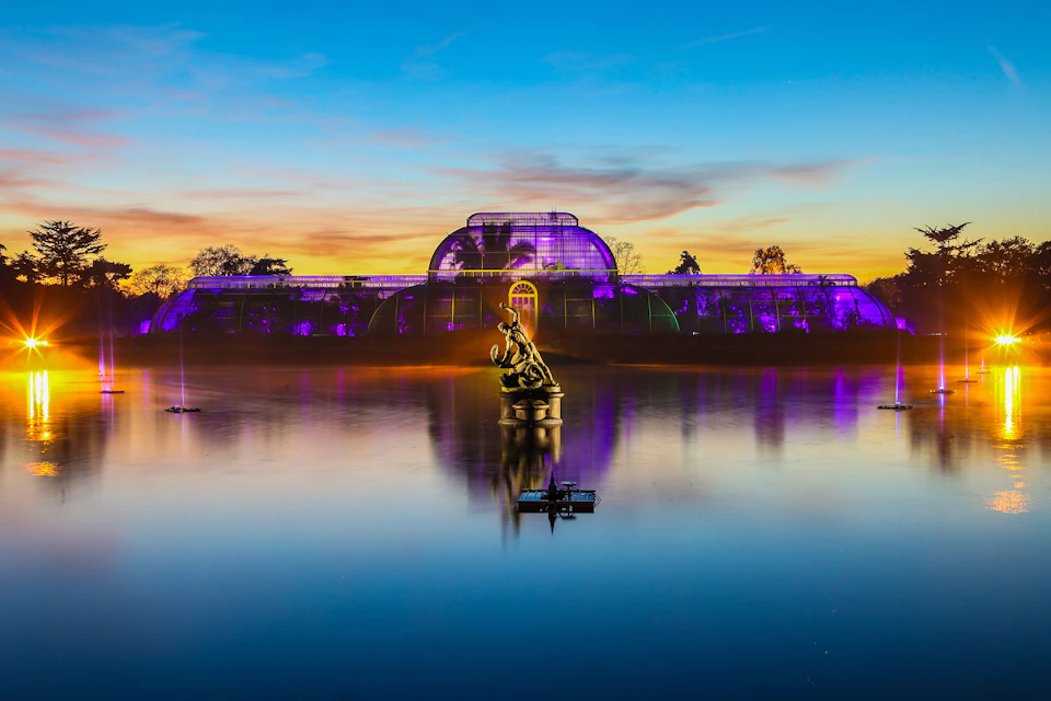 A purple-lit glasshouse with a statue in front, reflected in a tranquil lake at sunset with a boat in the foreground.