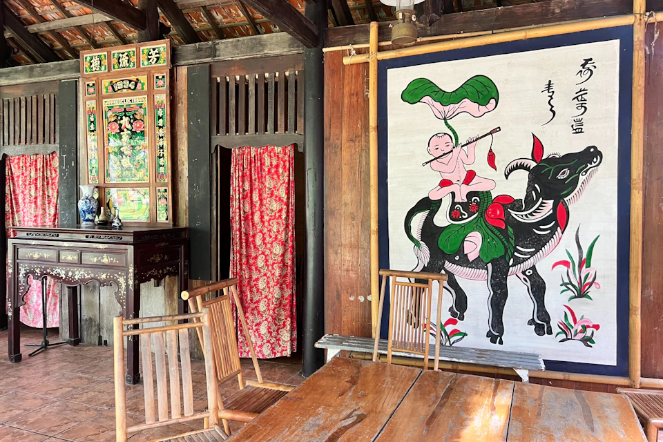 A traditional wooden room with a red floral curtain, wooden chairs around a table, and a wall hanging depicting a child on a black bull.