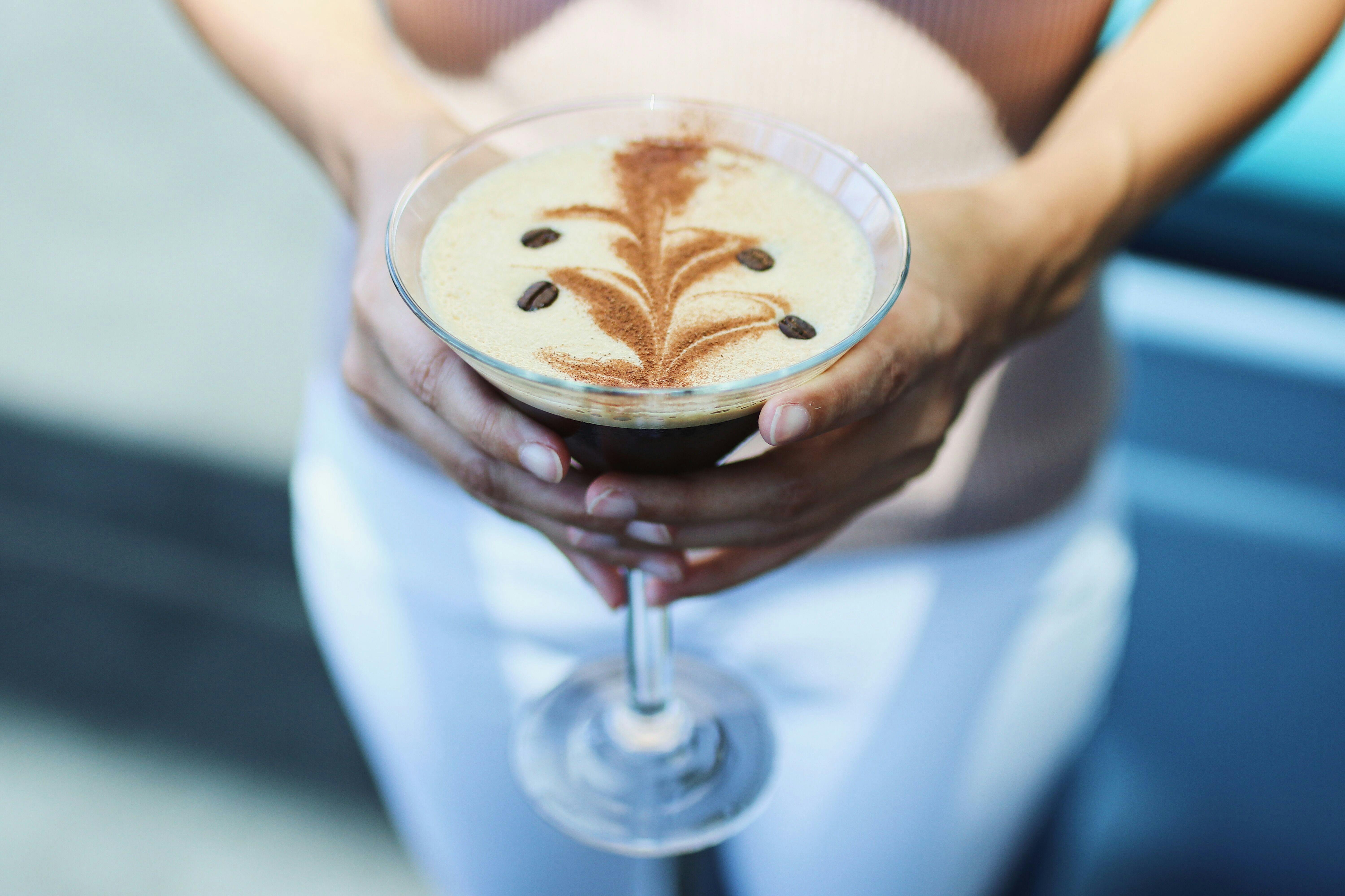 A person holding a martini glass with a creamy drink, topped with latte art and coffee beans.