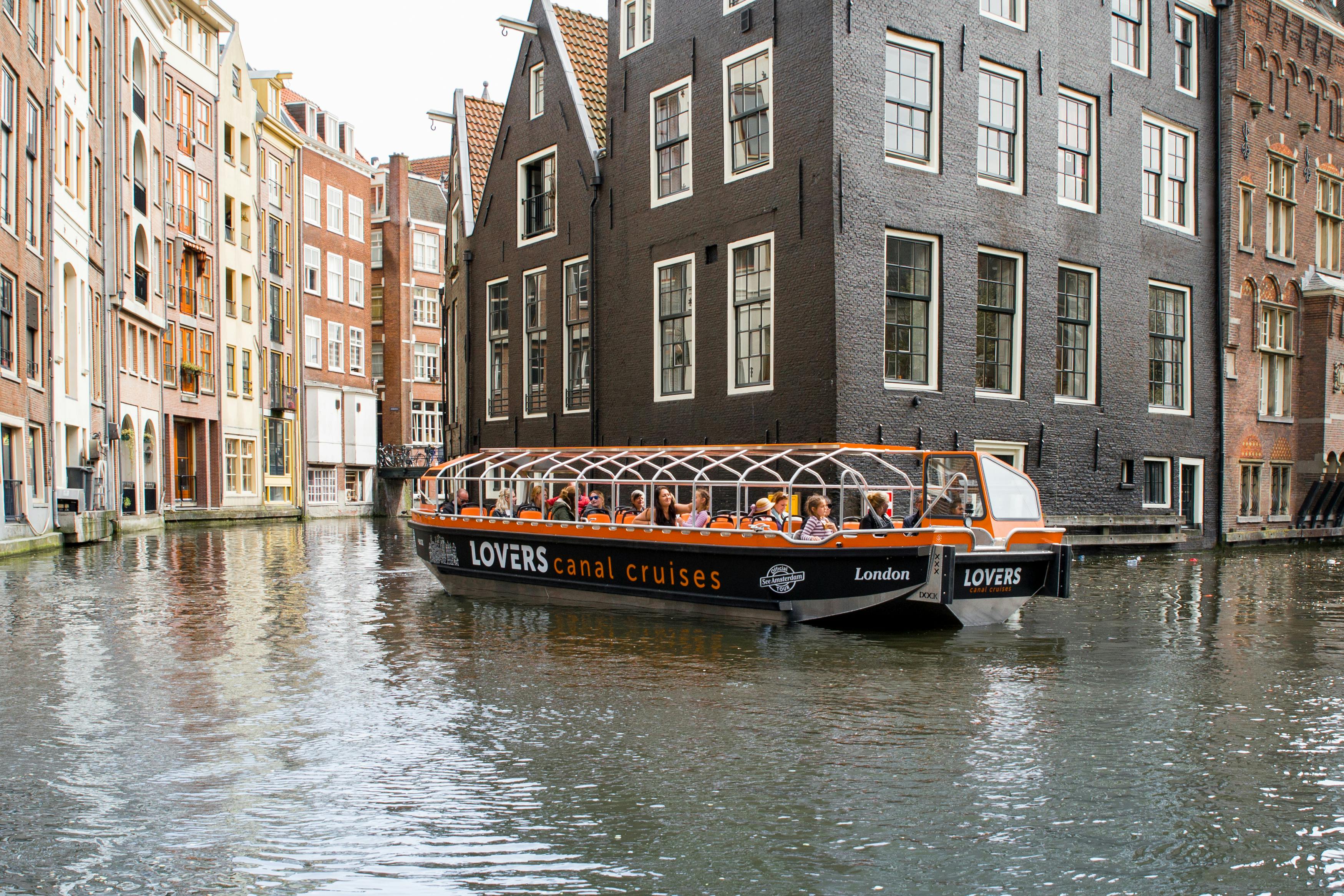 Boot met het label "LOVERS rondvaart over de grachten" met passagiers, bij donkere bakstenen gebouwen langs een gracht in een Europese stad.