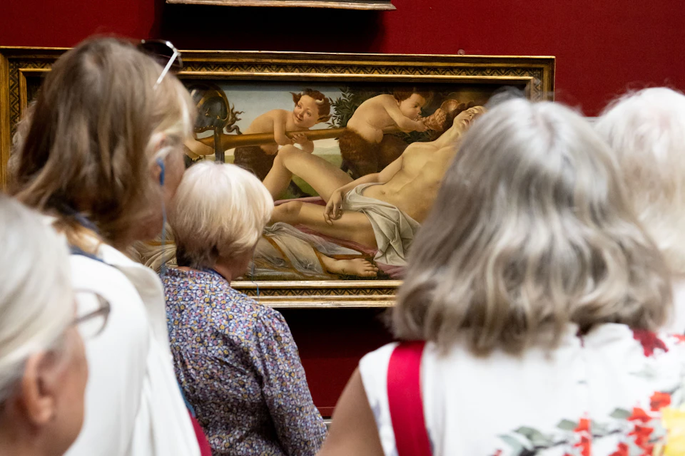 People viewing a classical painting depicting a reclining figure and cherubs, displayed in a red-walled gallery.