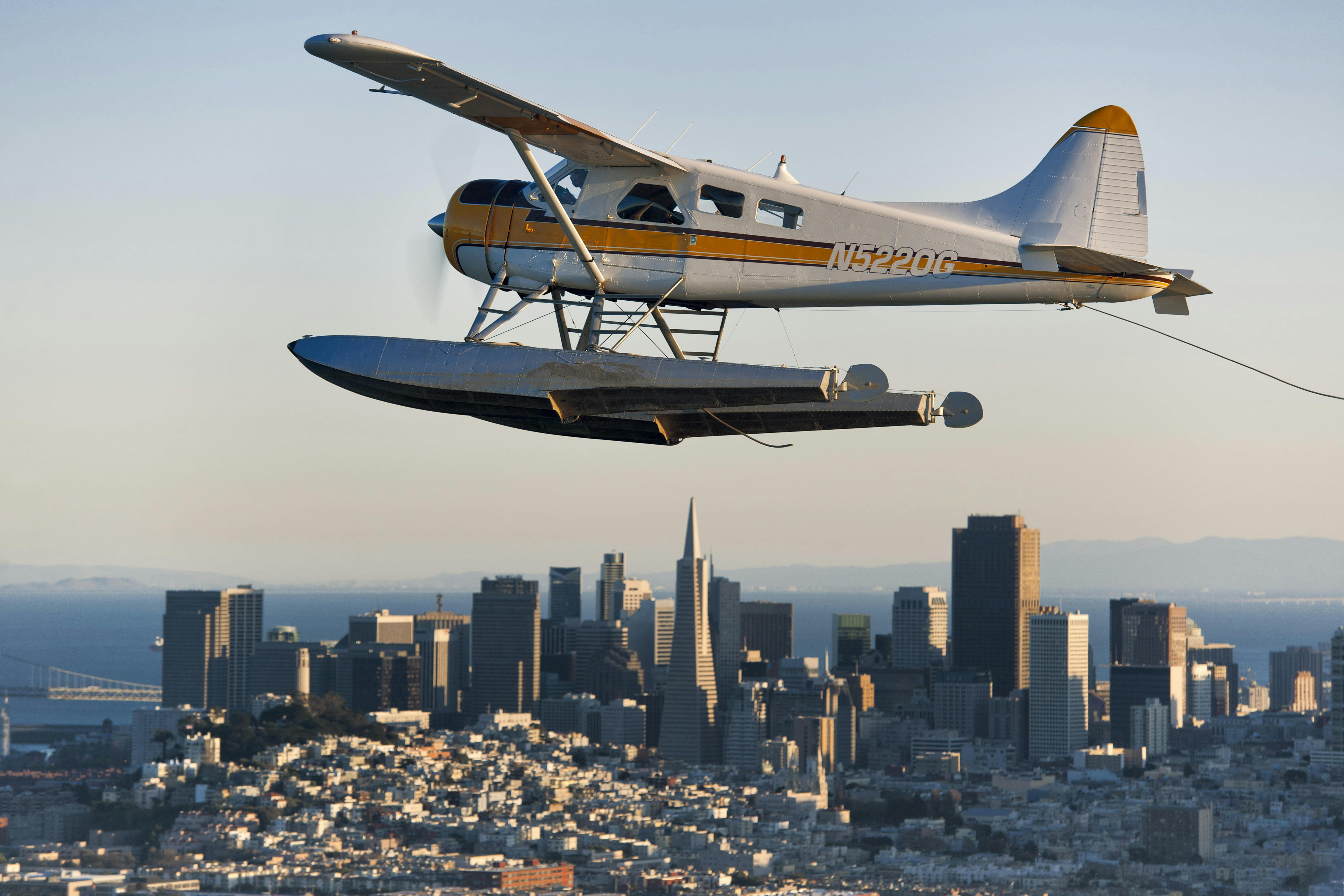 A seaplane flying over a city skyline with tall buildings and the ocean in the background.