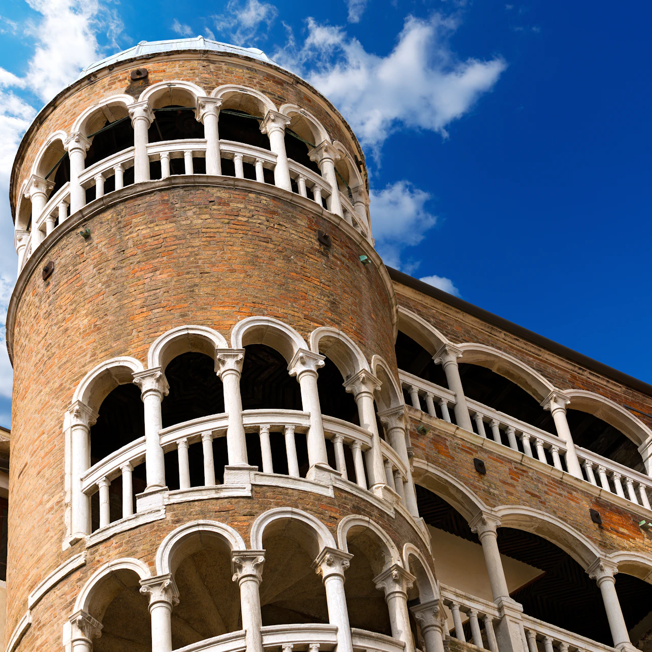 A round brick tower with multiple levels of white arches against a bright blue sky with scattered clouds.