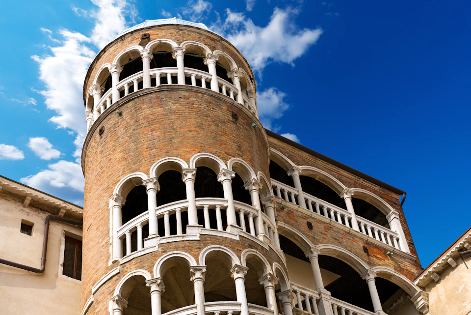 A round brick tower with multiple levels of white arches against a bright blue sky with scattered clouds.