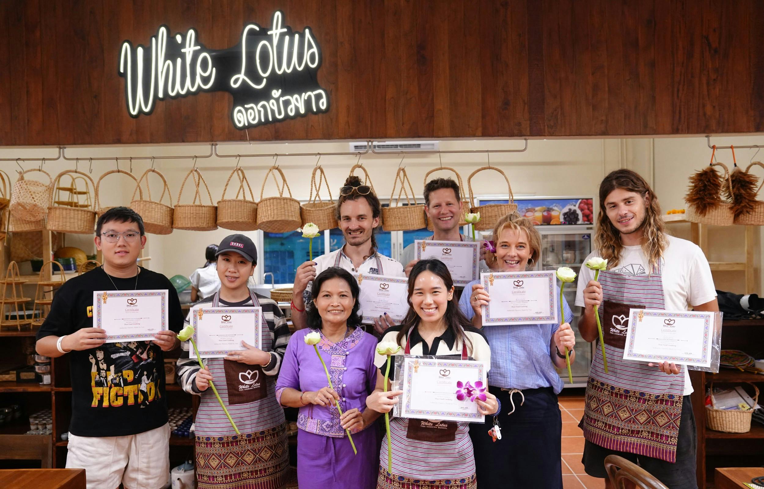 Seven people posing indoors, holding certificates and flowers, in front of a sign reading "Taste Lotus" in a cooking setting.